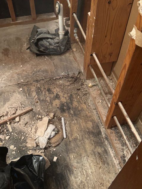 Bathroom remodel in progress: exposed plumbing, debris on damaged wooden floor.