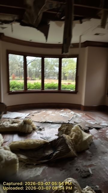 A room with a bay window and dark trim, featuring debris and insulation scattered across the floor and a damaged ceiling.