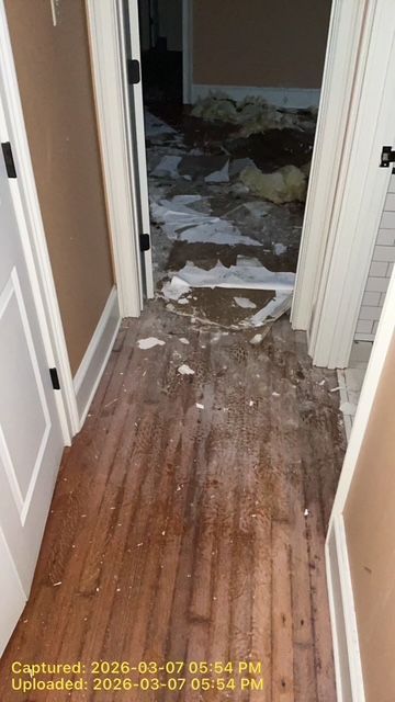 A narrow hallway with hardwood floors covered in white debris, leading into a room filled with scattered construction waste.