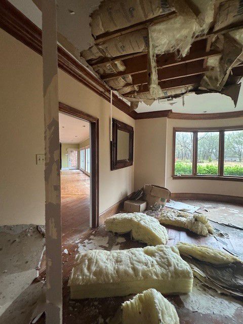 A room under renovation with exposed ceiling joists, hanging insulation, and debris scattered across the floor.