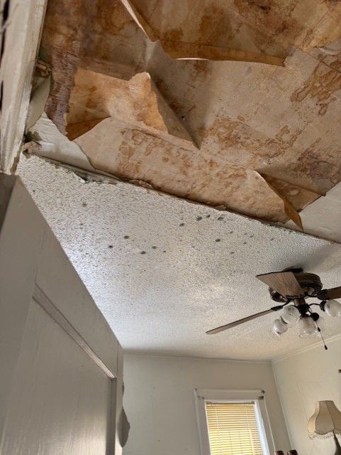 Damaged ceiling with exposed brown material and peeling white popcorn texture in a room with a ceiling fan.