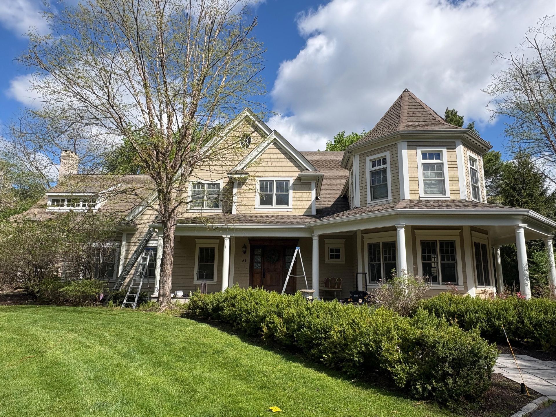 Large beige house with a turret, white porch, and green lawn under a cloudy sky