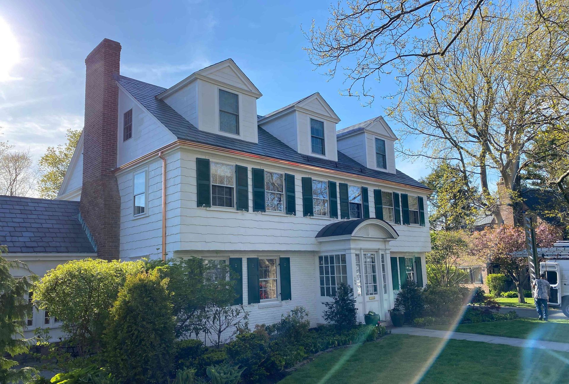 White two-story house with green shutters, brick chimney, and front porch in a sunny yard