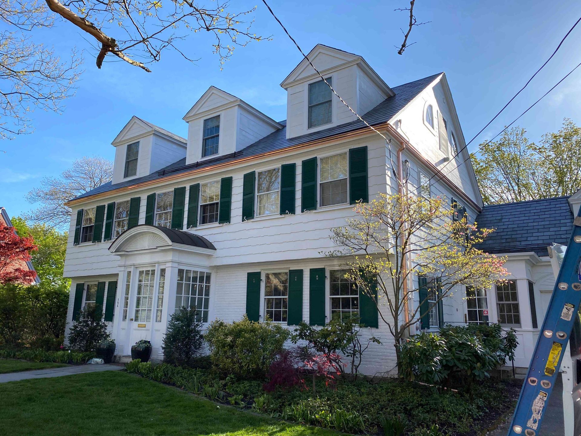 White colonial-style house with green shutters, dormer windows, and a ladder in the front yard under a blue sky