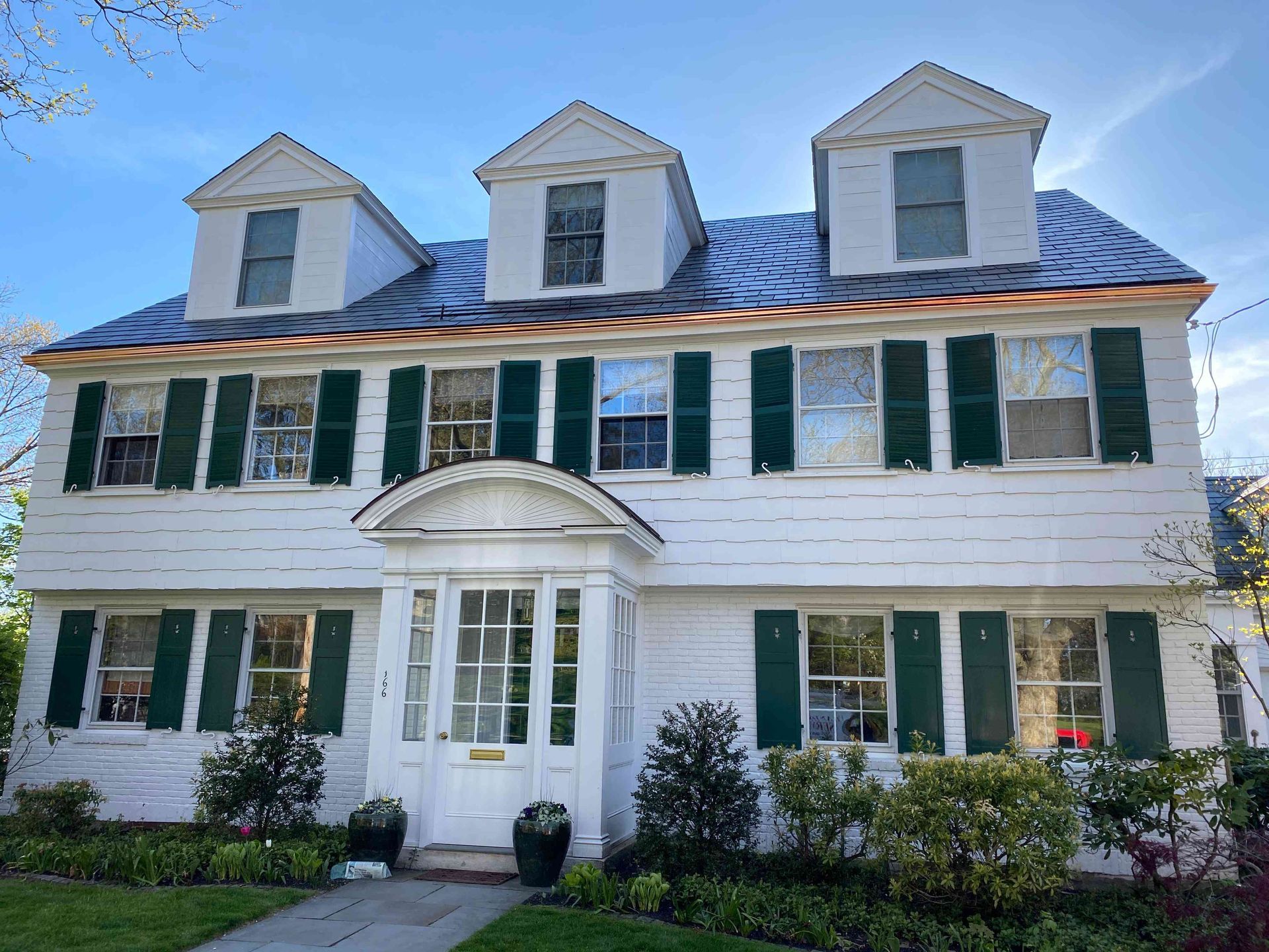 Two-story white house with green shutters, blue roof, and dormer windows under a partly cloudy sky