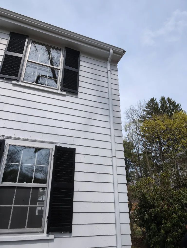 White house exterior with two windows and black shutters, viewed from below against a cloudy sky.