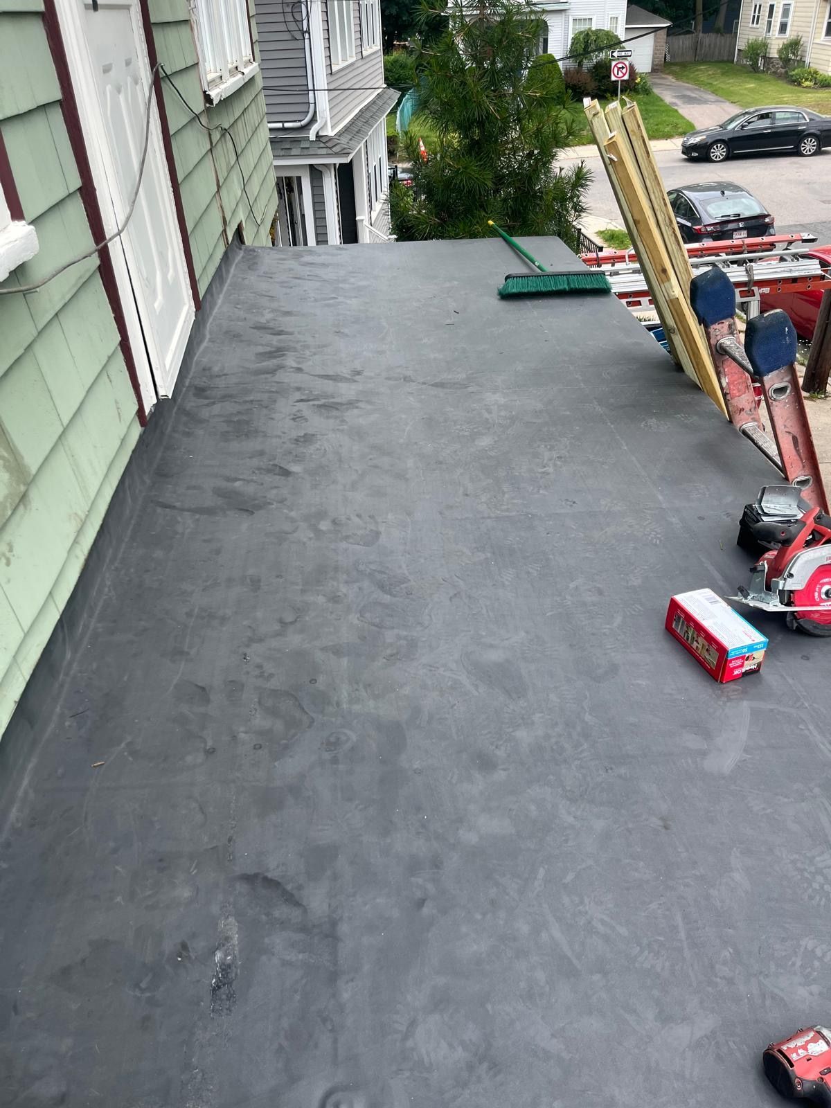 Flat rooftop with gray waterproofing beside a house, overlooking a street and parked cars below