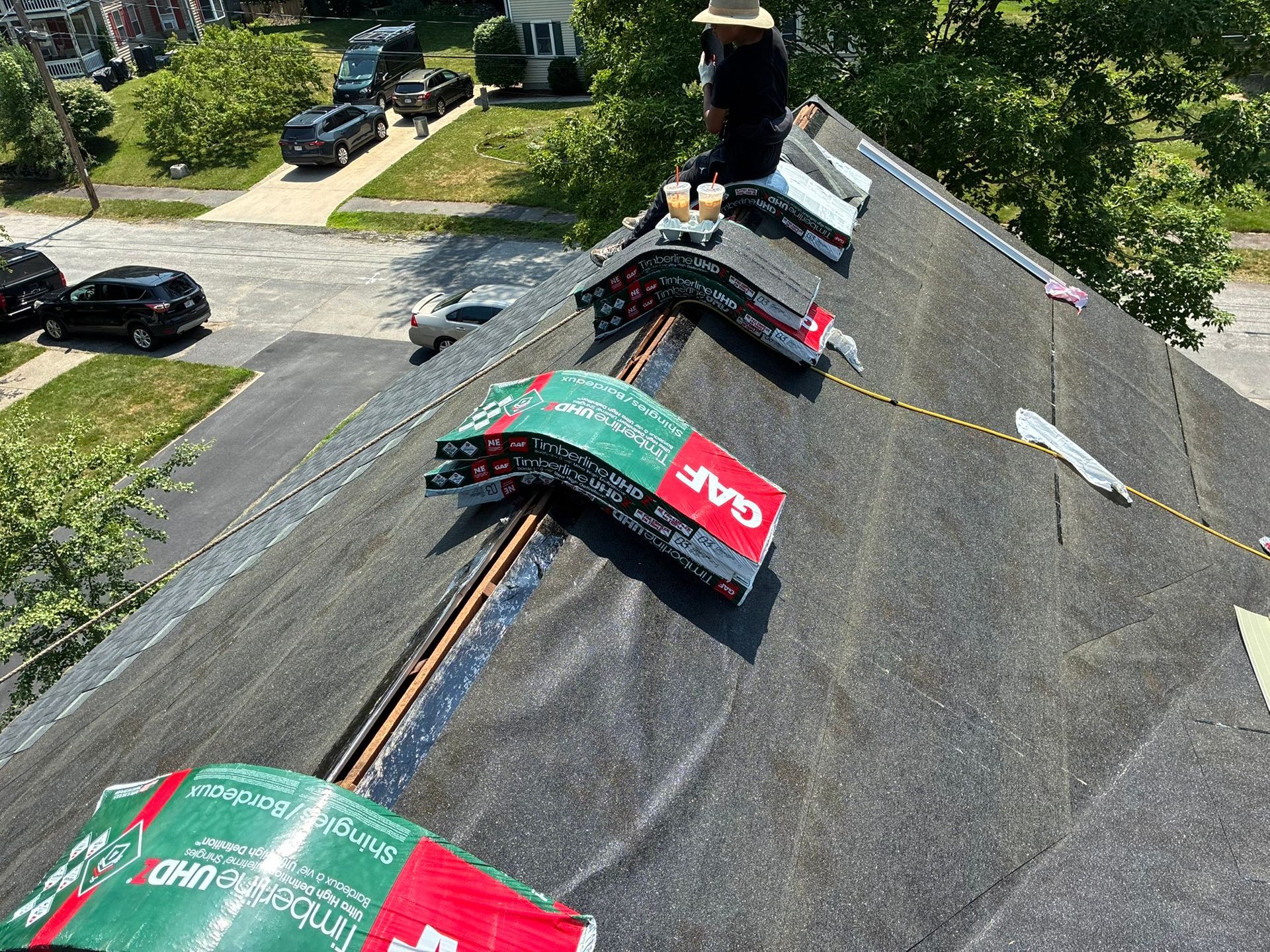 Roofers installing green and red shingles on a steep gray roof, with a residential street below.
