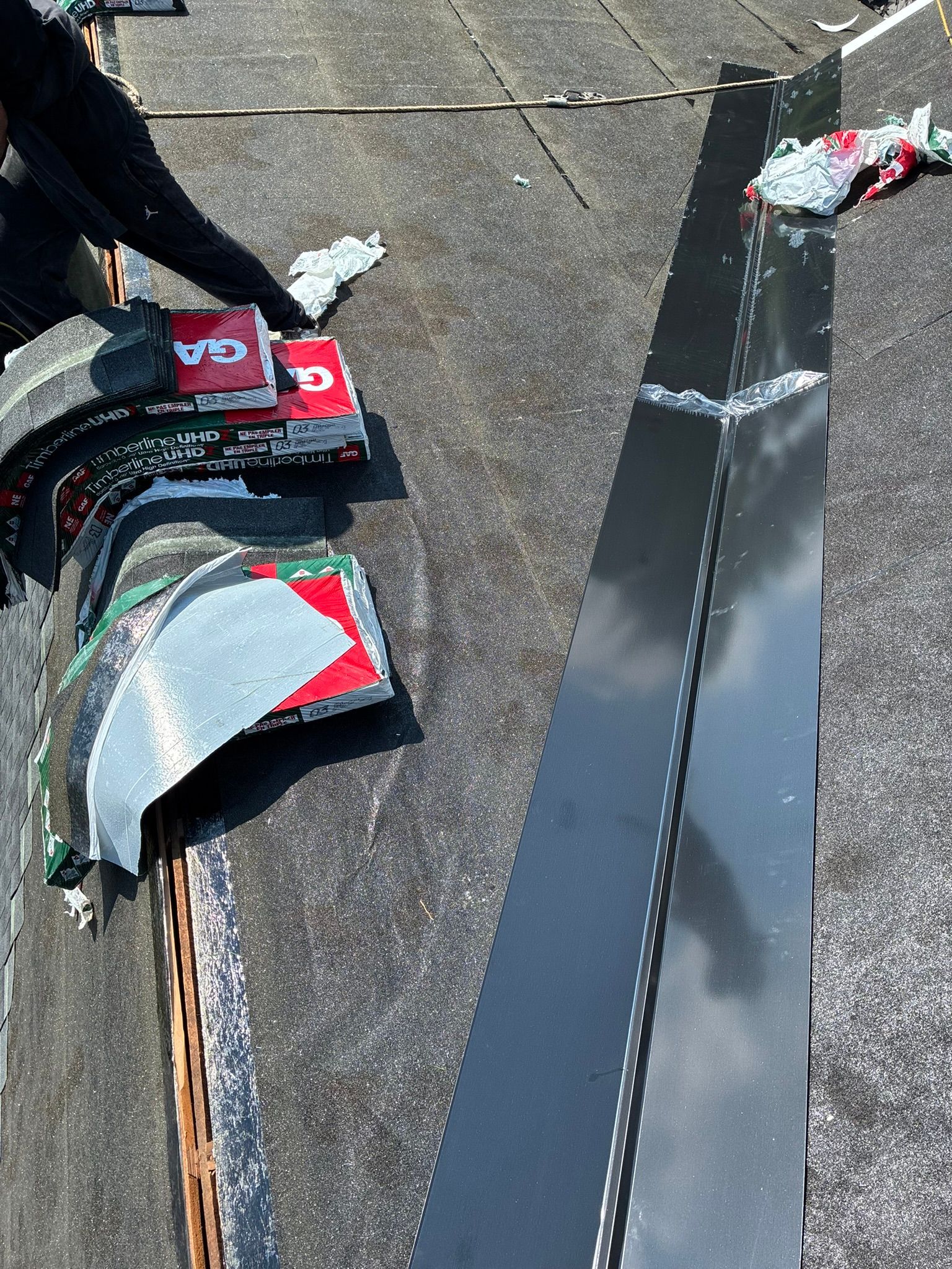 Roofing materials and a person’s hand beside a black metal panel on a shingled roof