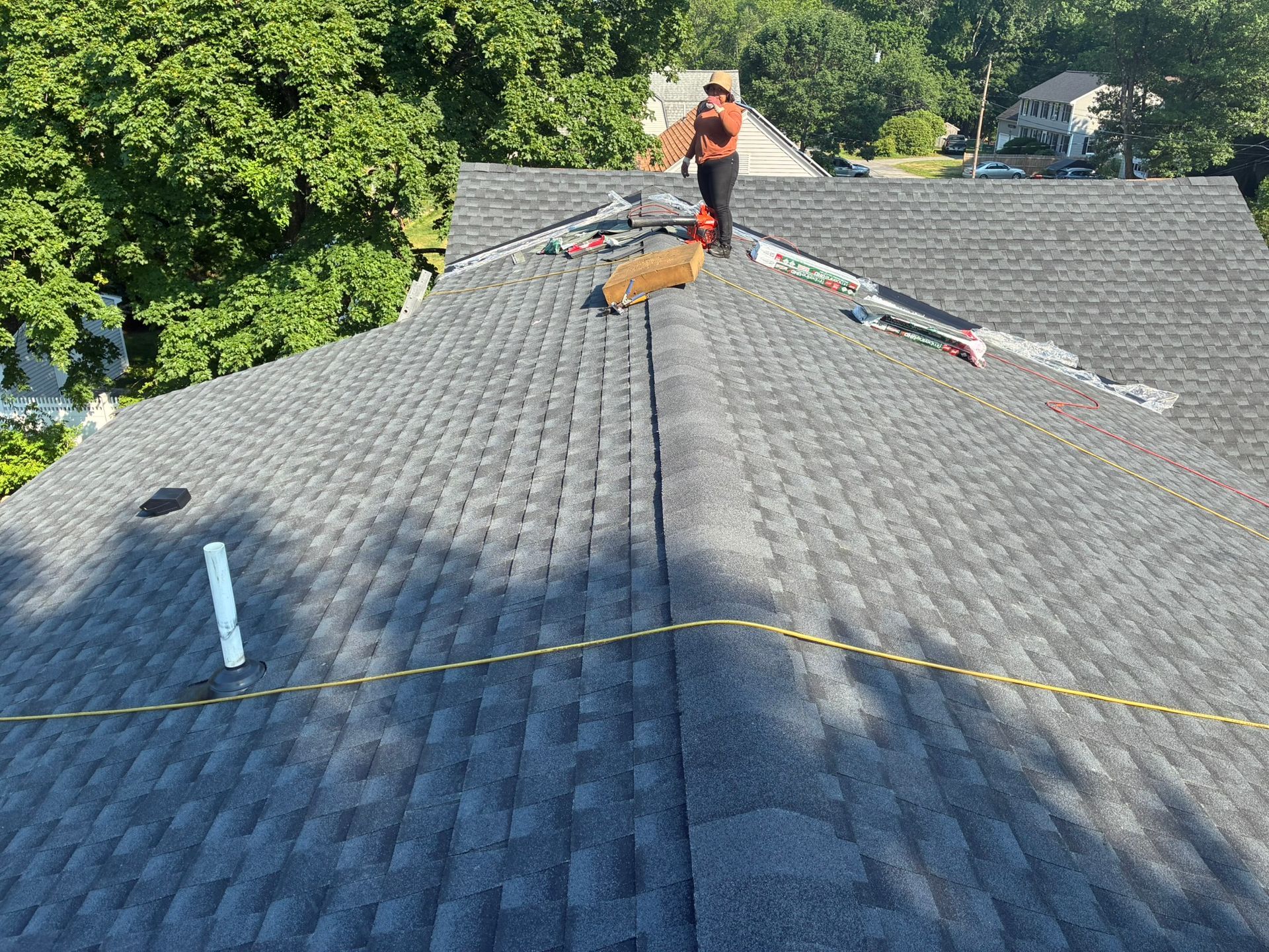 Roofing worker on a shingled roof, near a ridge line with safety ropes and trees in the background
