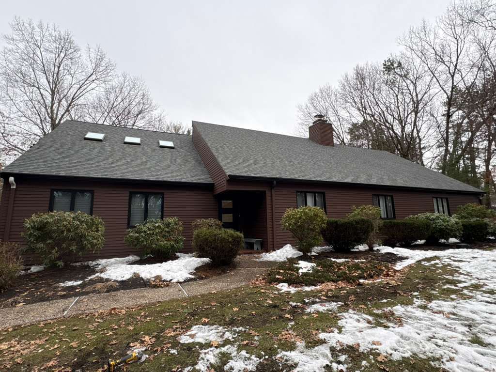 Brown house with dormer windows and a front walkway, surrounded by leafless trees and patchy snow