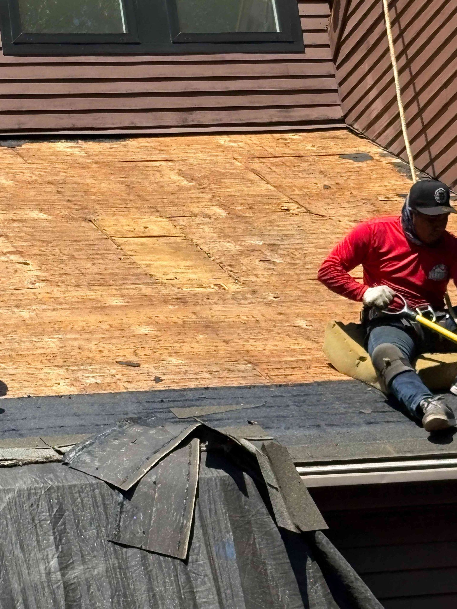 Worker in red shirt repairing a roof with exposed plywood and shingles.