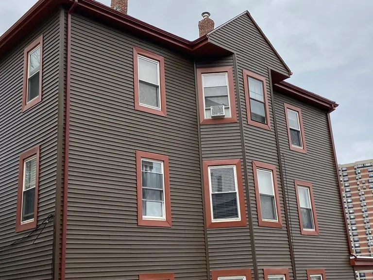 Three-story brown house with red-trimmed windows and a sloped roof under an overcast sky