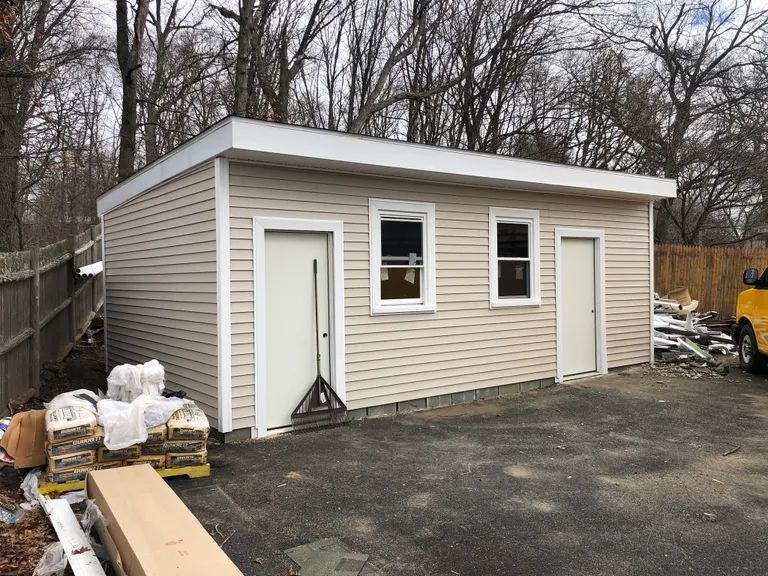 Small tan garage with two white doors, set on a paved lot beside a fence and leafless trees.