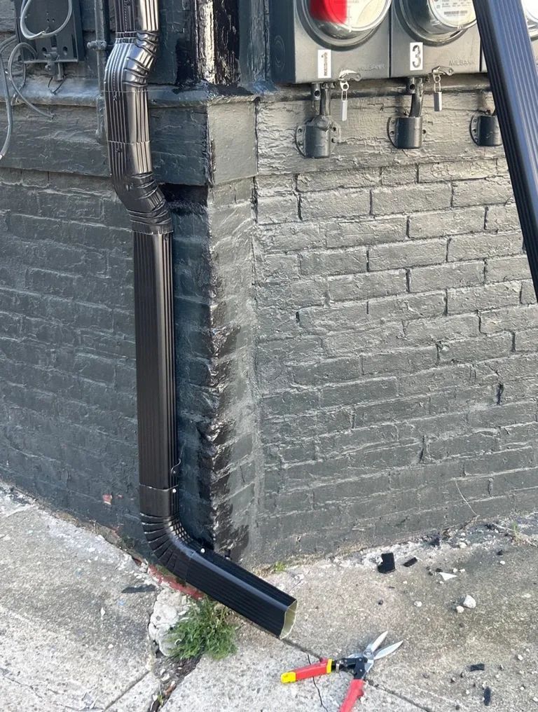 Black brick building corner with a downspout and utility pipes near the sidewalk, with a broom and debris below