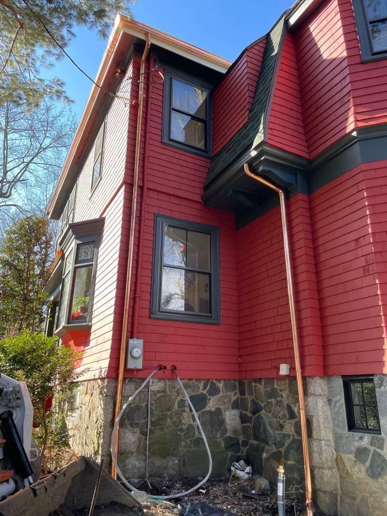 Red house exterior with stone foundation, black-trim windows, and a downspout on a sunny day