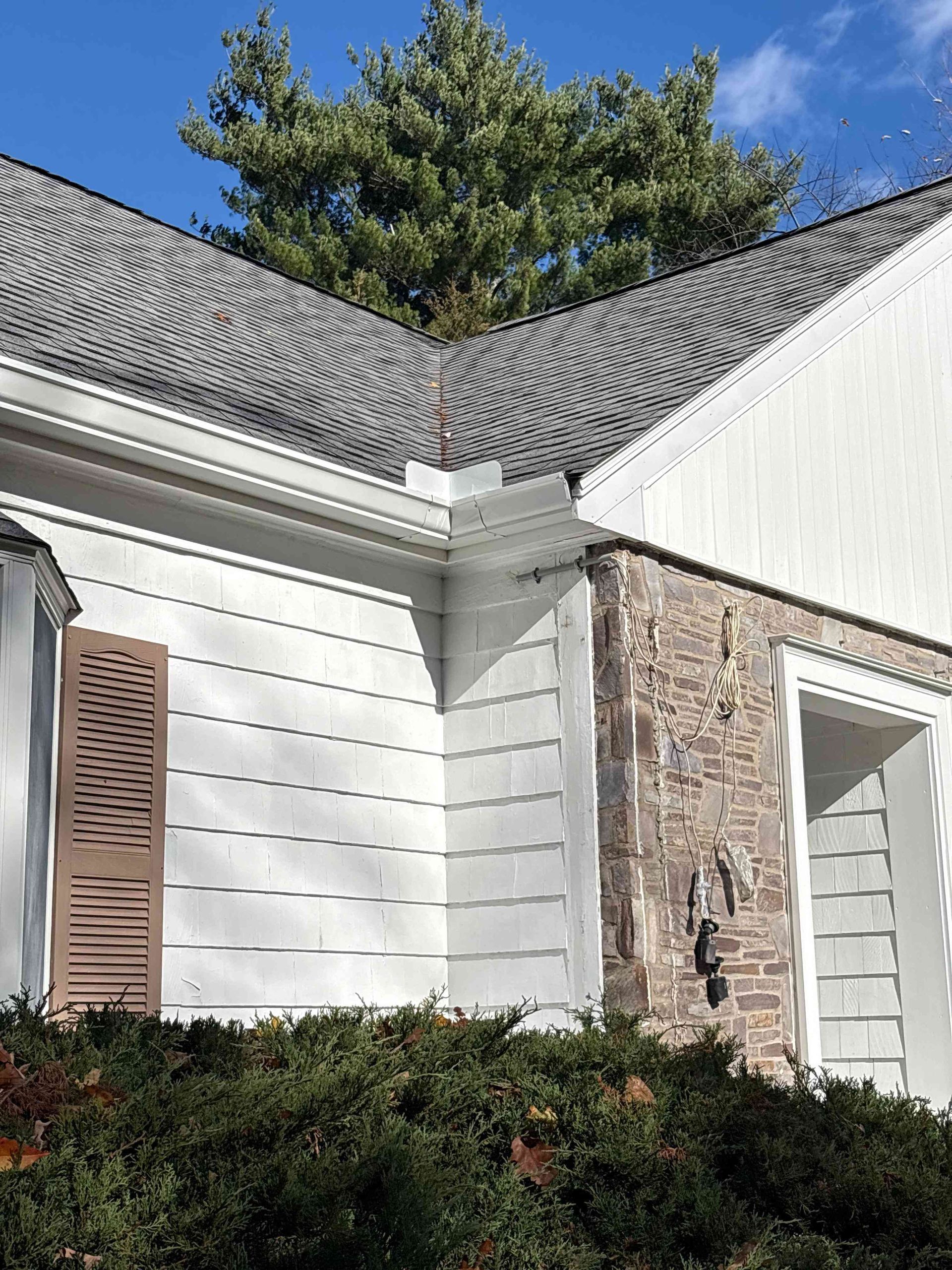 White house exterior with stone and siding, dark shingle roof, and shrubs in front under a blue sky