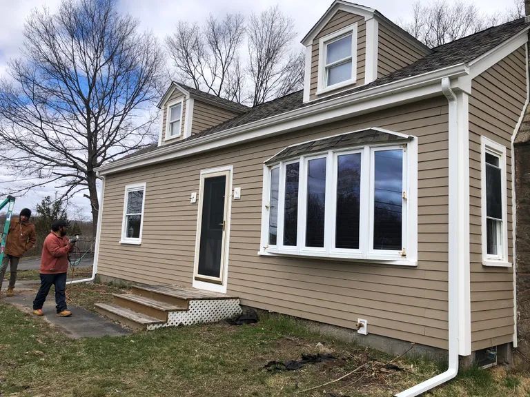 Small tan house with white trim and a large bay window, seen from the yard with a person standing nearby.