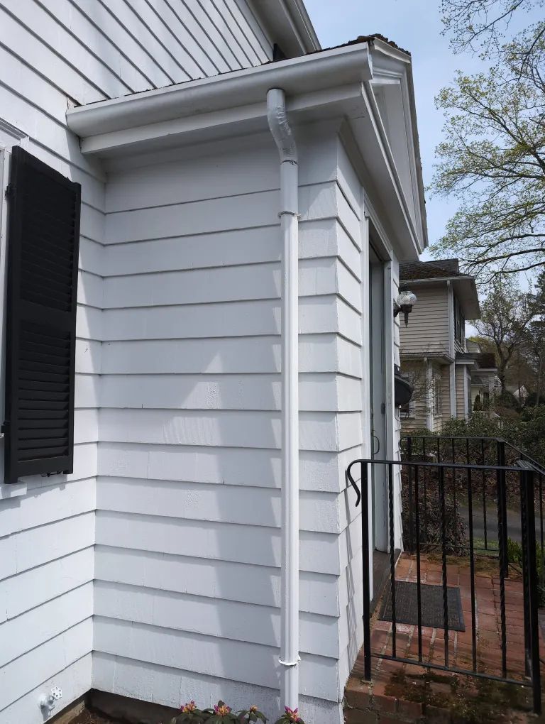 White house corner with gray siding, black shutters, and a downspout beside a small fenced brick walkway
