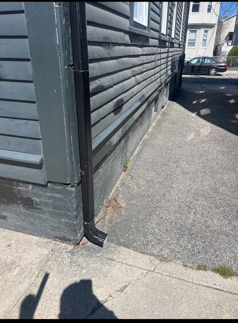 Side of a gray wooden building beside a narrow paved walkway, with a black drainpipe and parked car in the background