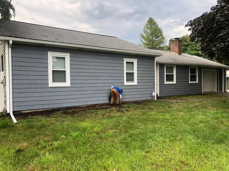 Person working near the blue-gray side of a house on a grassy lawn