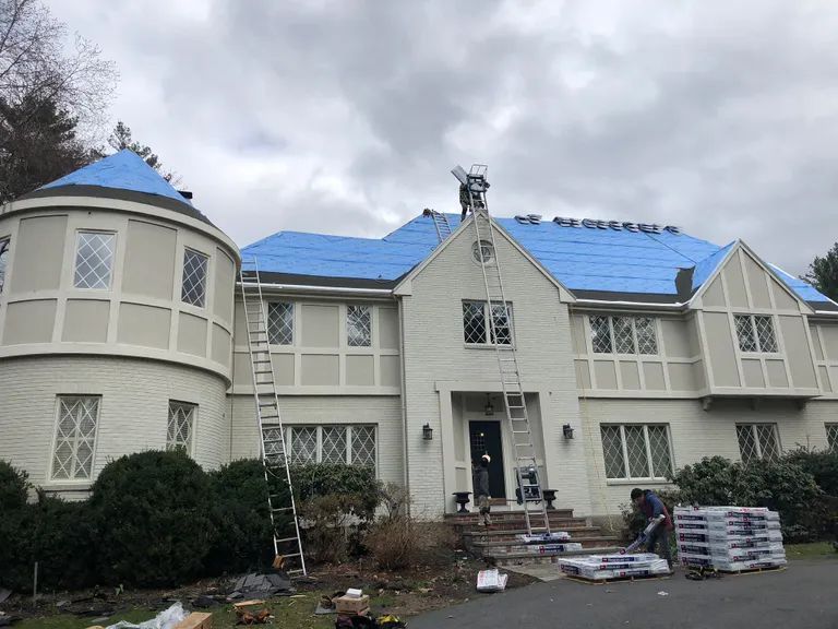 Large two-story house under renovation, with blue roof sheathing and construction materials in front.