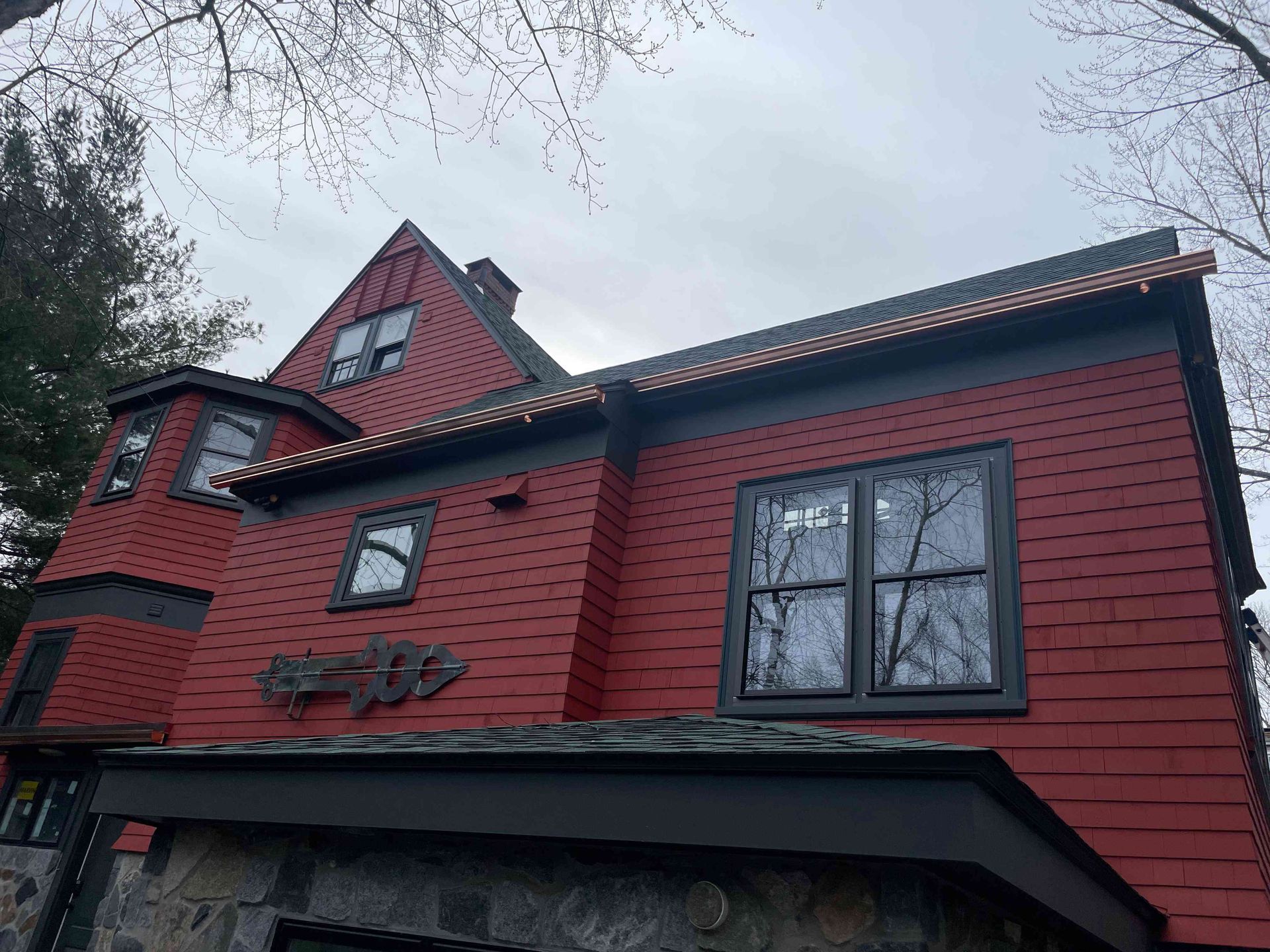 Red house with dark trim and steep roof, viewed from below through bare tree branches