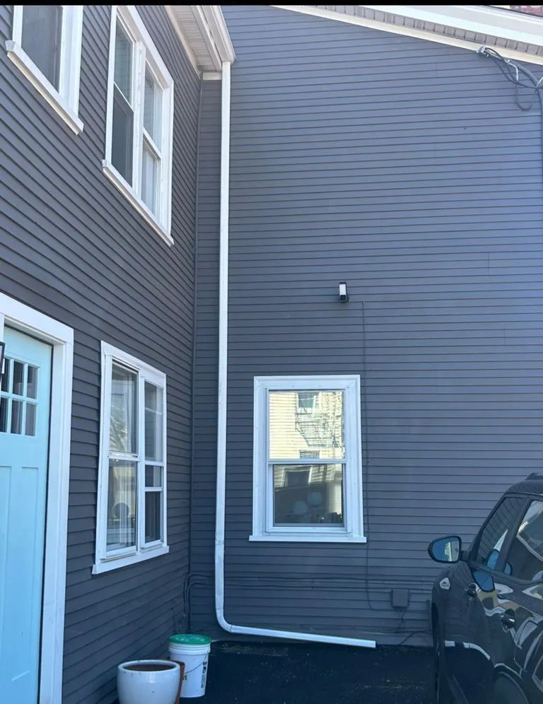 Side of a gray house with windows, downspout, and a parked black vehicle in a narrow driveway.