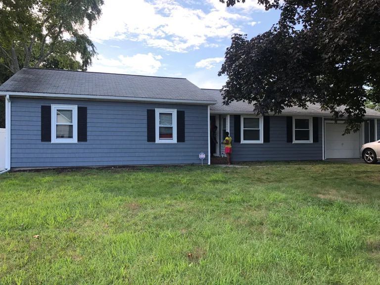 Single-story blue house with white trim, front lawn, trees, and a person standing by the doorway.