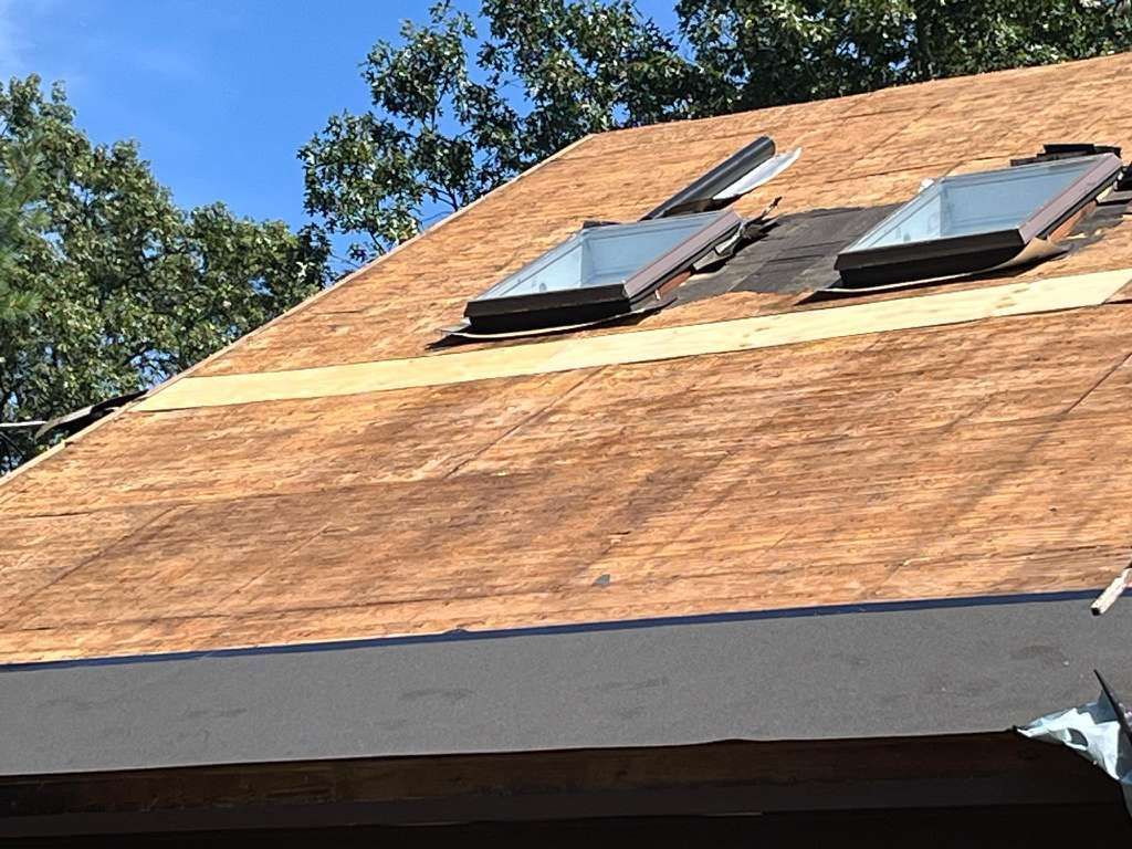 Roof with brown shingles and two skylights under a blue sky, with trees in the background