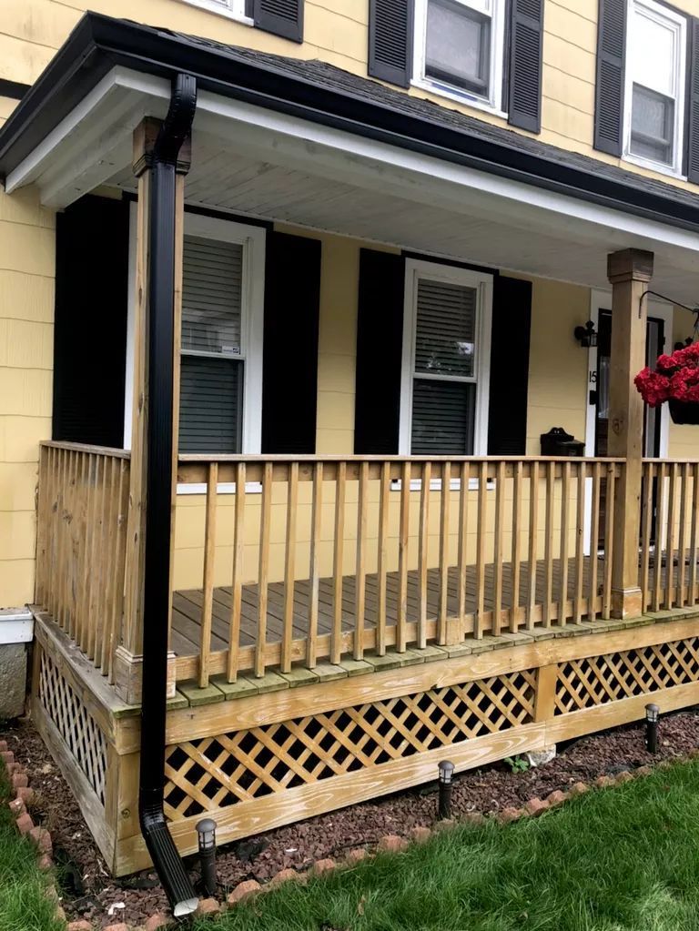 Front porch with beige siding, black shutters, and a wooden railing in front of a house.