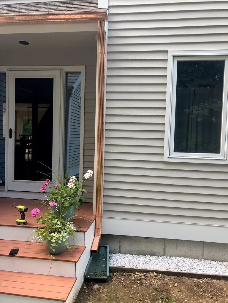 Front porch of a house with potted flowers, a black door, and beige siding beside a window.
