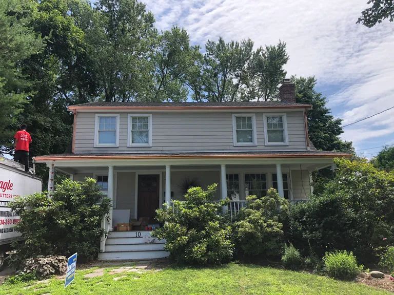 Two-story house with a front porch, beige siding, and overgrown bushes under a cloudy sky