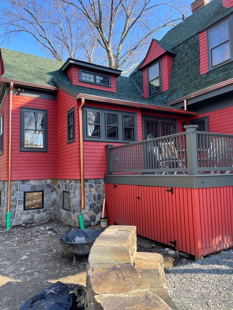 Red house with stone foundation and gray deck railing in a wooded yard