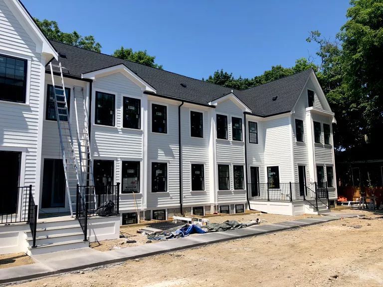 Row of white townhouses under construction with black-trim windows and a dirt driveway under a blue sky