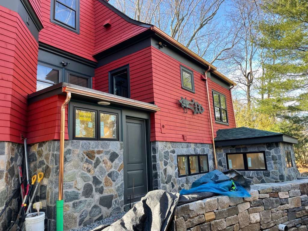 Red house with stone base and black trim, viewed from the side in a wooded yard.