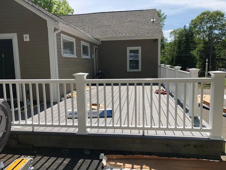 Wooden deck with white railing attached to a gray house on a sunny day.