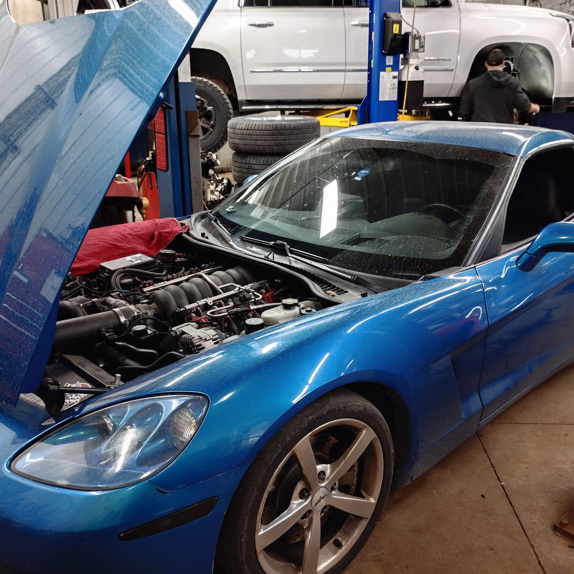 Blue sports car with hood open in a garage, mechanic working nearby and a truck in the background
