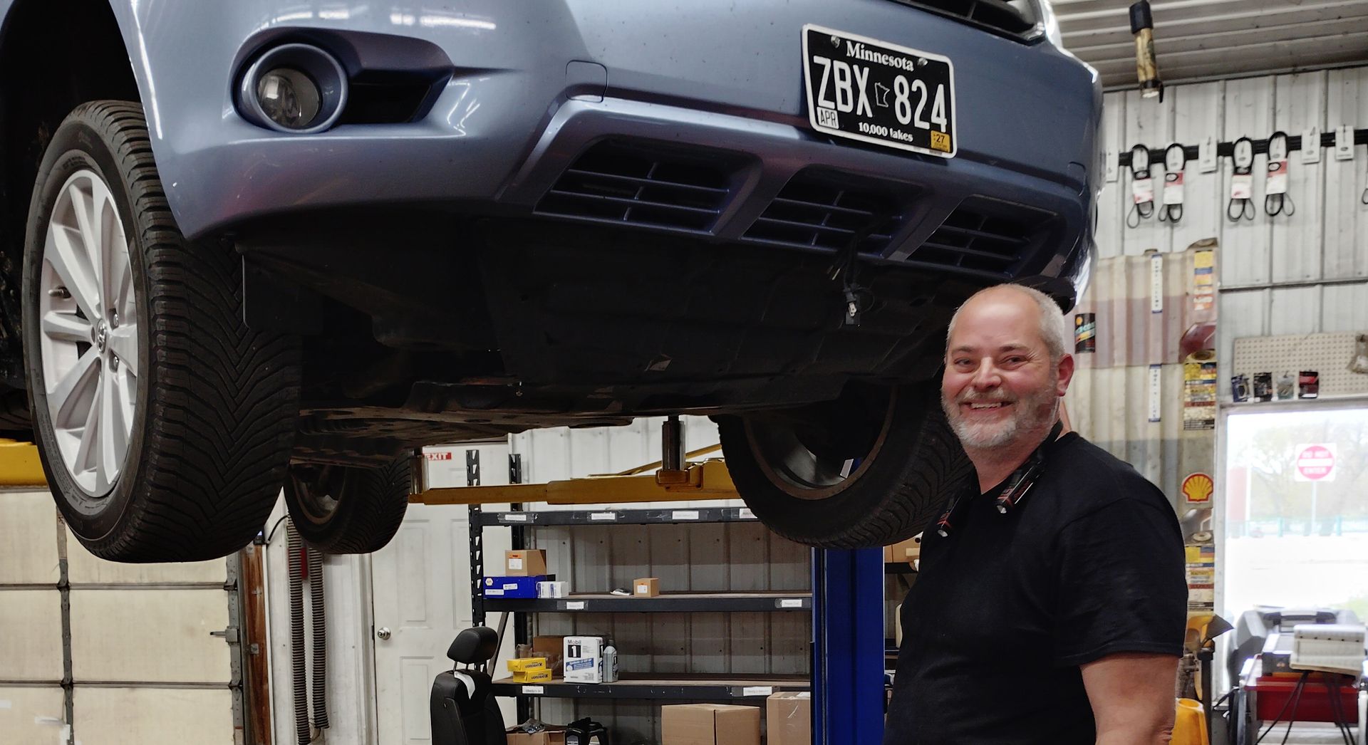 Gray car lifted on a shop hoist, with a smiling mechanic standing underneath in an auto garage.