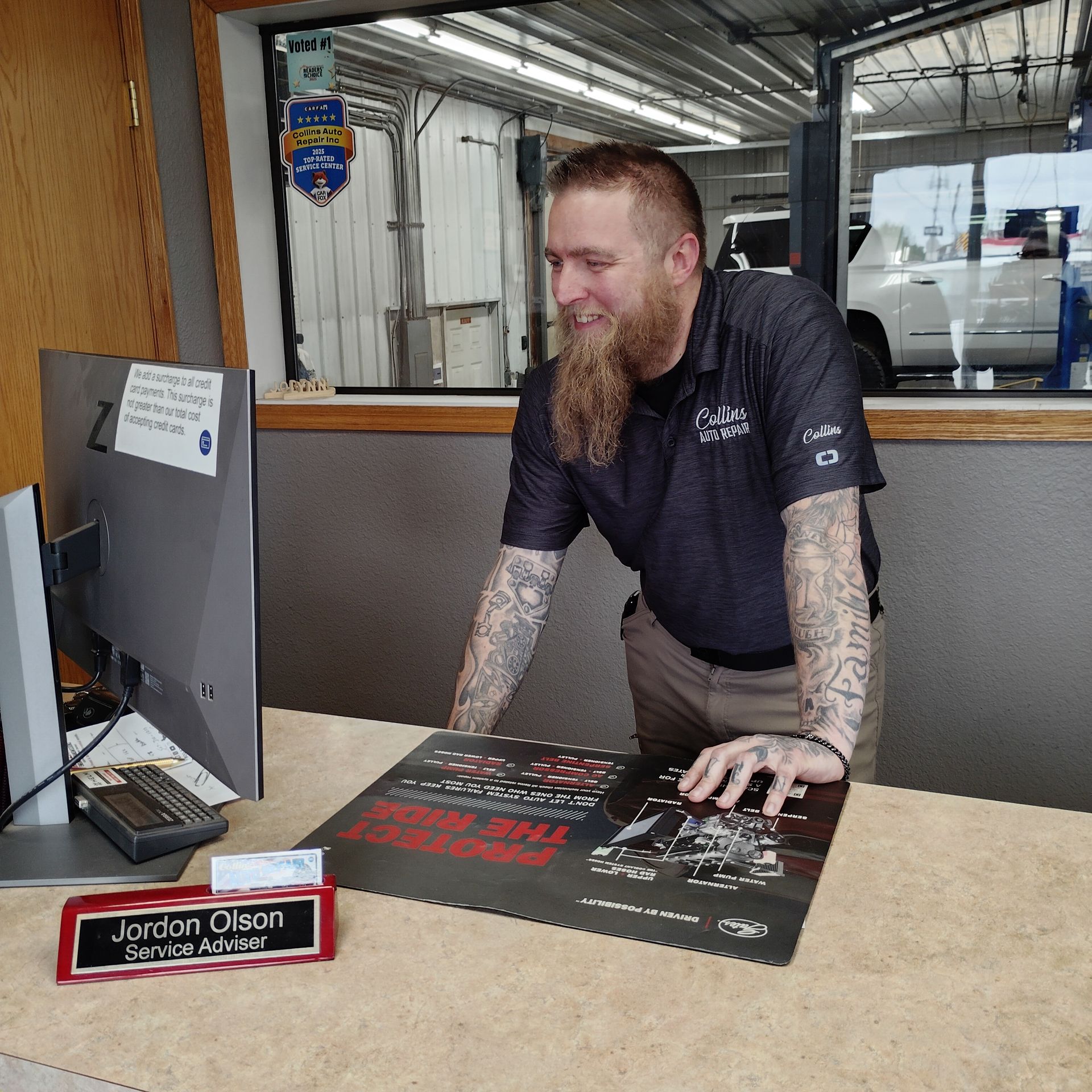 Mechanic smiling at a desk beside a monitor, in a garage office with a nameplate and brochure.