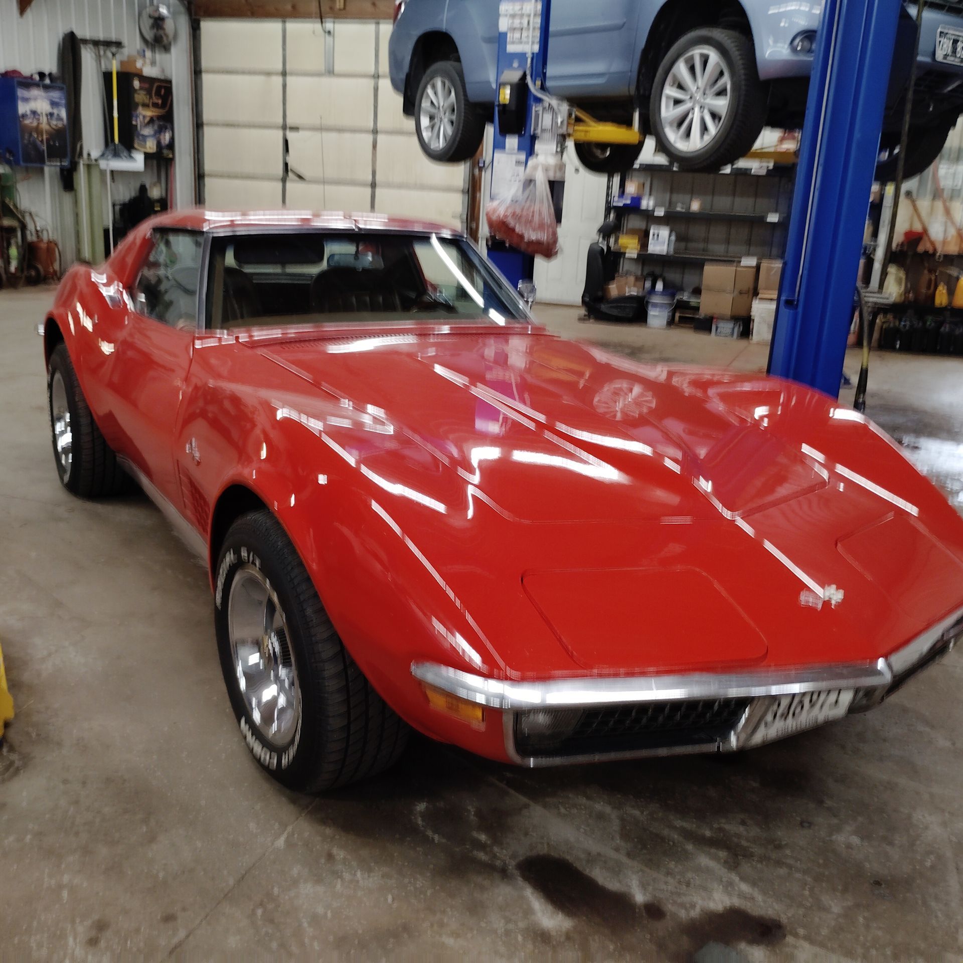 Red classic sports car in a garage, with a blue SUV lifted on a rack in the background.