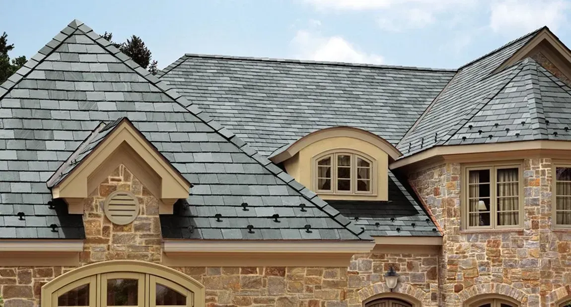 Stone house roof with gray slate shingles, dormer windows, and blue sky above