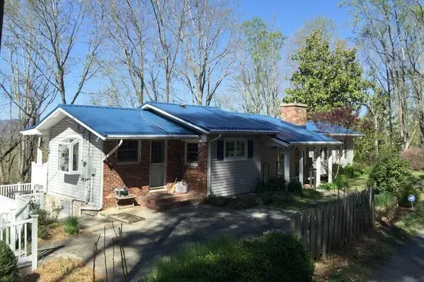 Single-story brick house with blue roof and chimney, surrounded by trees and a wooden fence