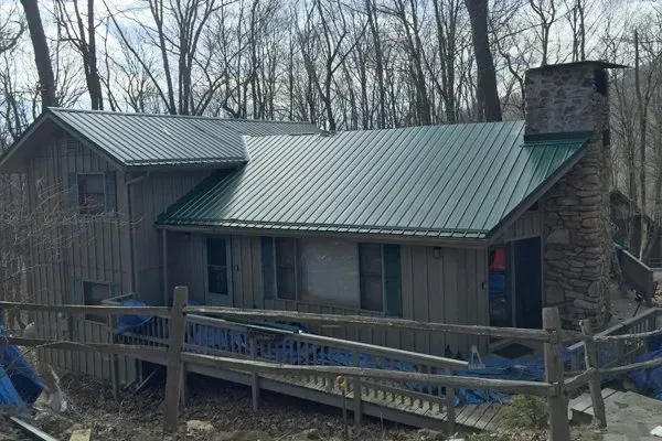 Rustic cabin with green metal roof and stone chimney in a wooded area, viewed from the front.
