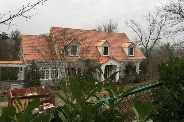 Two-story house with red roof and dormer windows, surrounded by trees and greenery