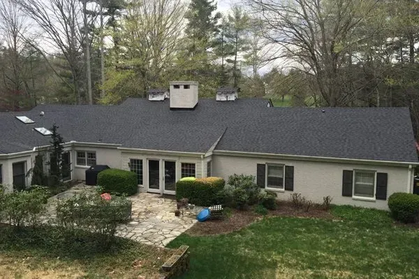 Single-story beige house with dark roof, white-trimmed windows, and a stone front path in a wooded yard