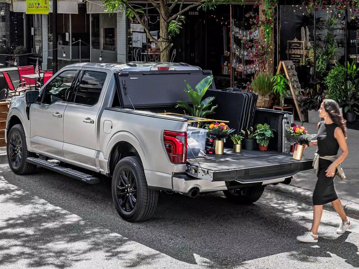 Silver pickup truck with flowers, woman removing flowers from the truck's bed, storefront background.