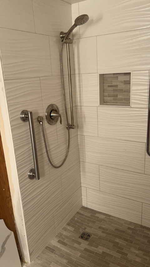 A tiled shower with a handrail, showerhead, and recessed shelf.