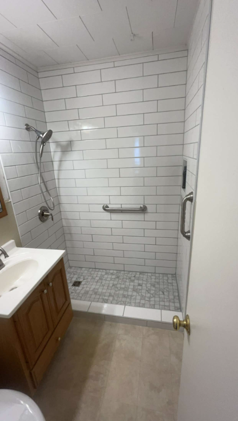 Shower with white subway tile, a grab bar, and a glass door; next to a vanity.