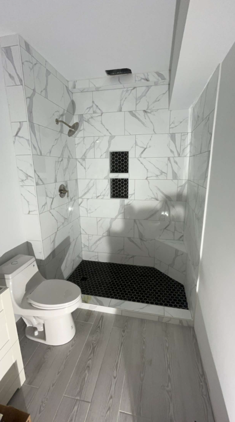 Bathroom with marble-look tiled shower, black mosaic floor, toilet, and gray wood-look floor.